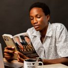 Young man reading Midas Touch book at desk, sitting with laptop and coffee mug.