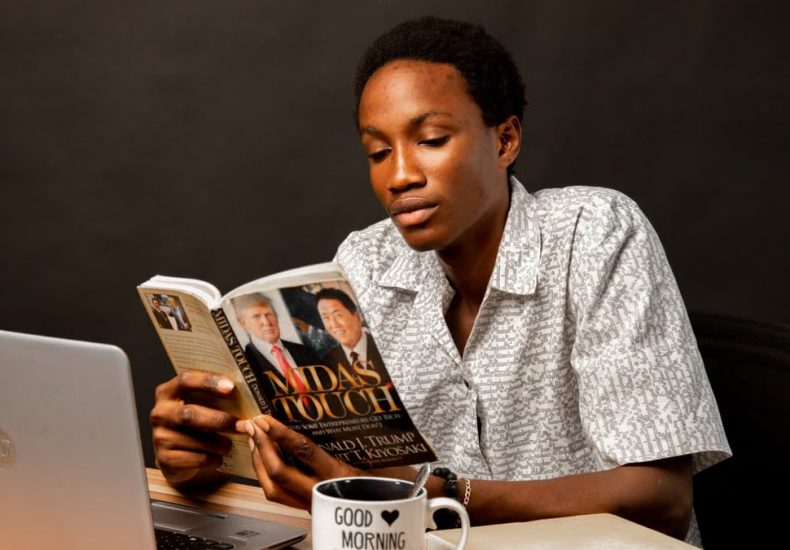 Young man reading Midas Touch book at desk, sitting with laptop and coffee mug.
