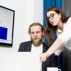Two colleagues analyzing business data on a laptop with a presentation screen in the background.