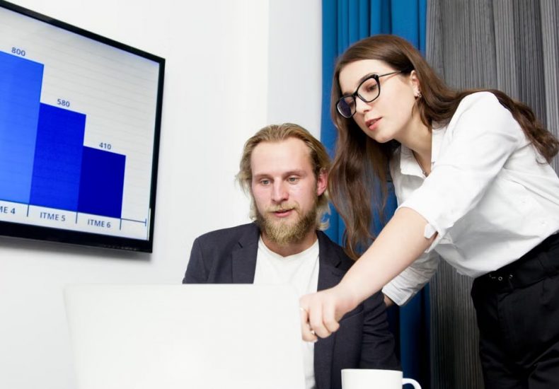 Two colleagues analyzing business data on a laptop with a presentation screen in the background.